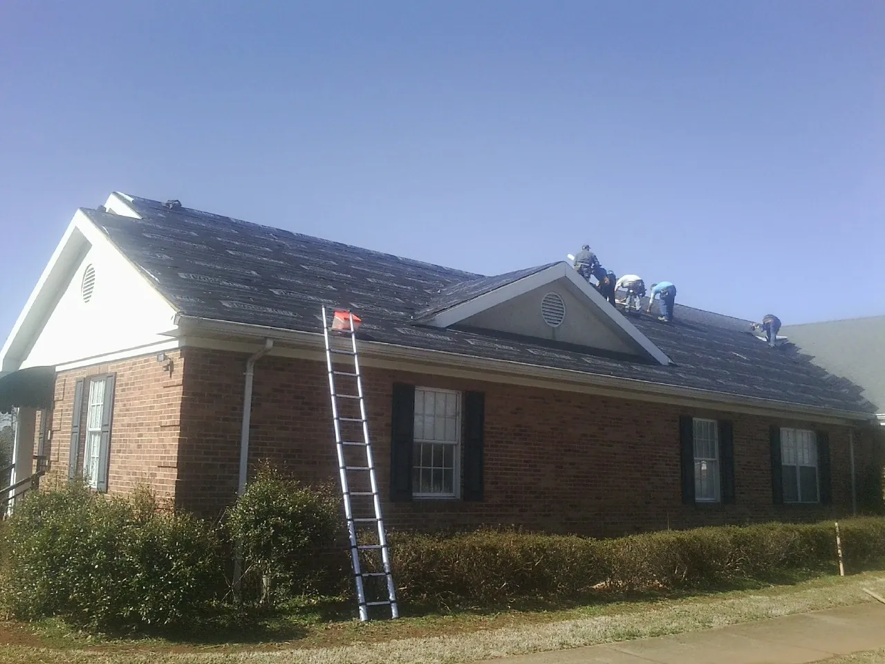 Crew tearing shingles off a brick ranch roof with a ladder set — Emergency Roof Repair work by our crew in Frankenmuth