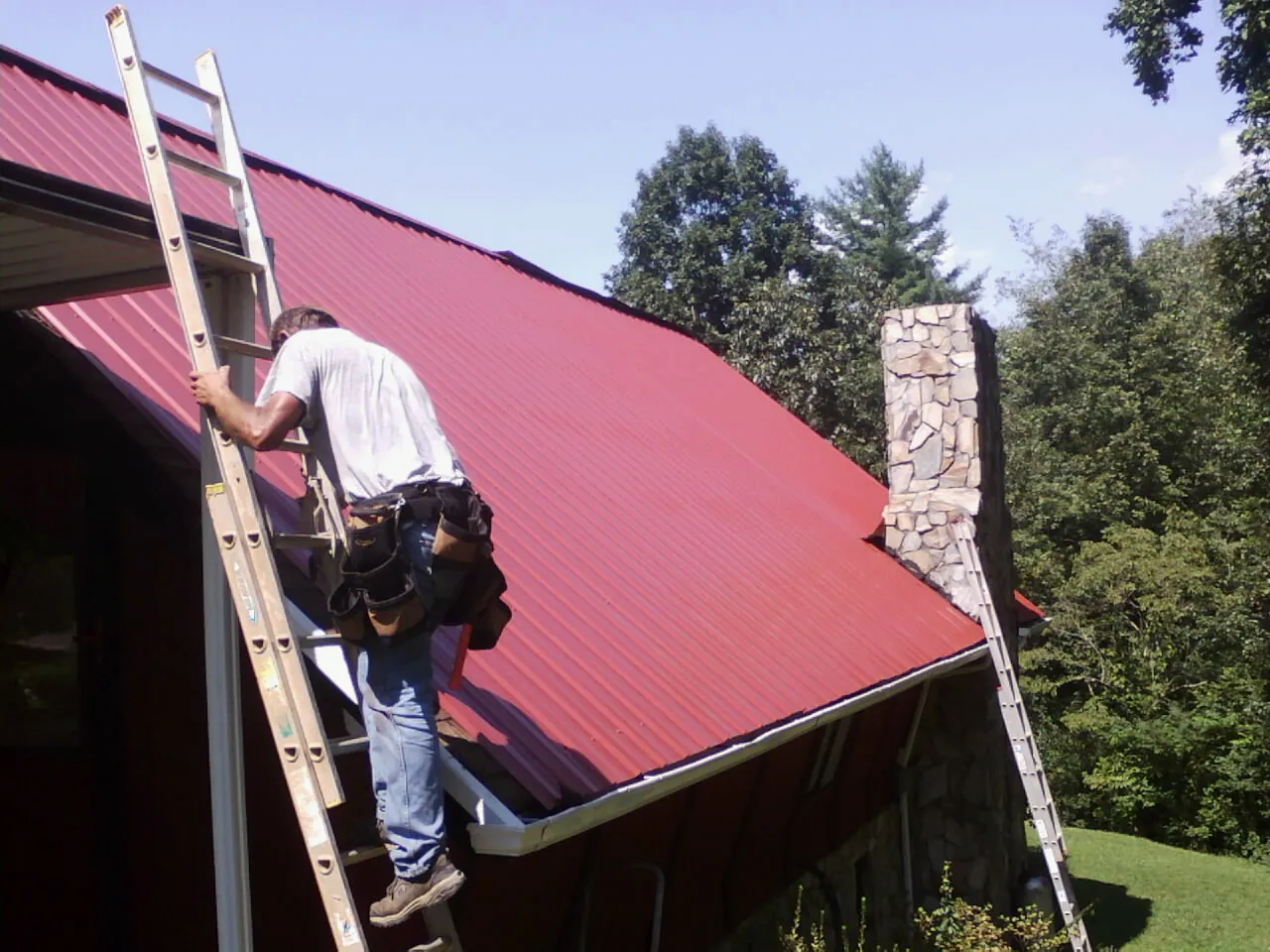 Roofer on a ladder beside a red metal roof and stone chimney — Soffit & Fascia Repair by Roofing Brothers in Frankenmuth